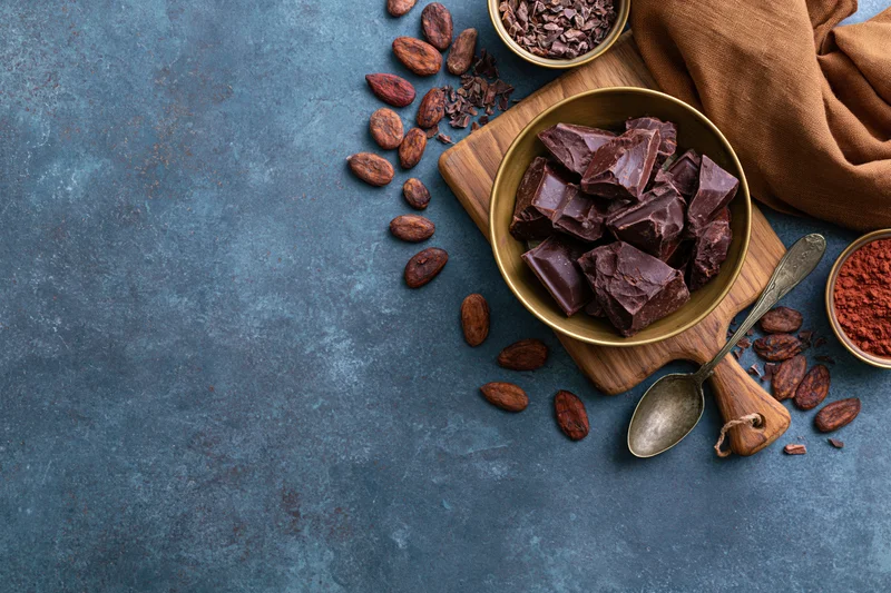 Dark chocolate chunks in a brass bowl on a wooden cutting board with cocoa beans and powder, arranged on a blue textured surface