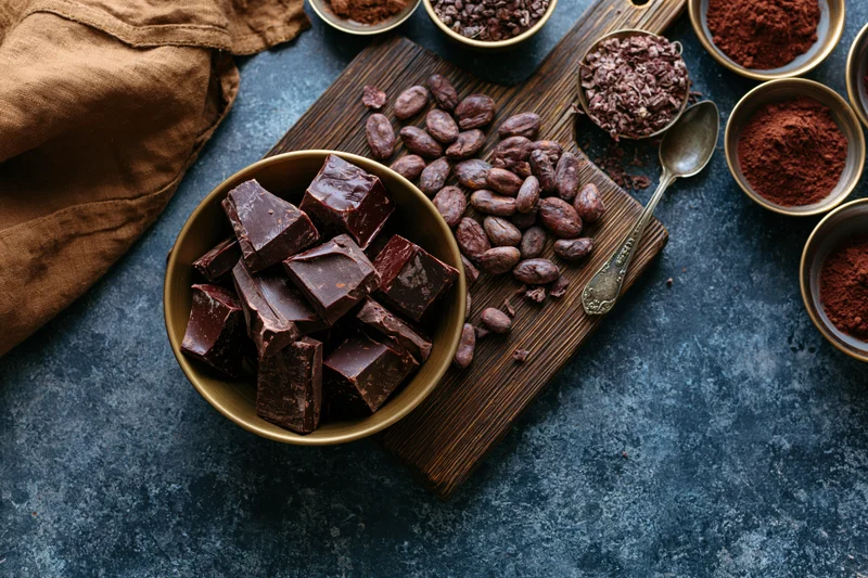 Dark chocolate chunks in a brass bowl with cocoa beans and powder on a wooden cutting board