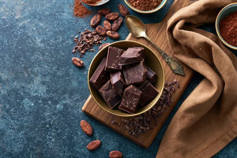 Dark chocolate chunks in a brass bowl on a wooden cutting board with cocoa beans and powder nearby