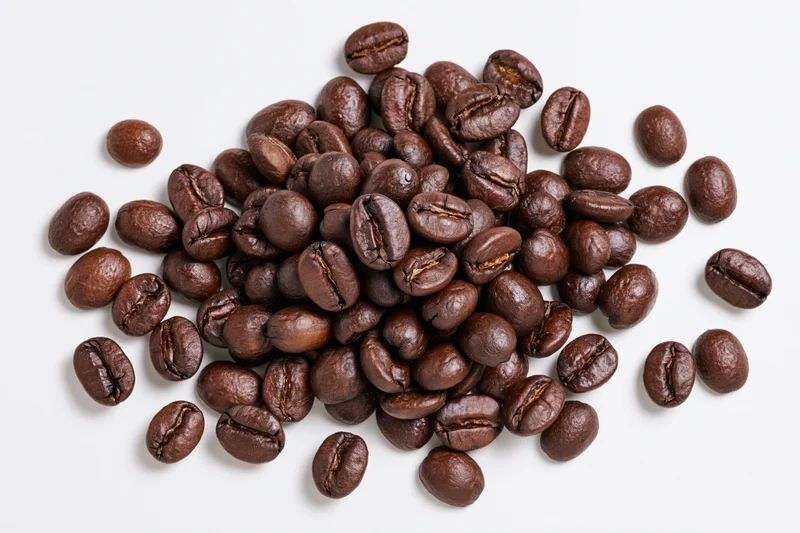 Dark roasted coffee beans scattered in a loose pile on a white surface