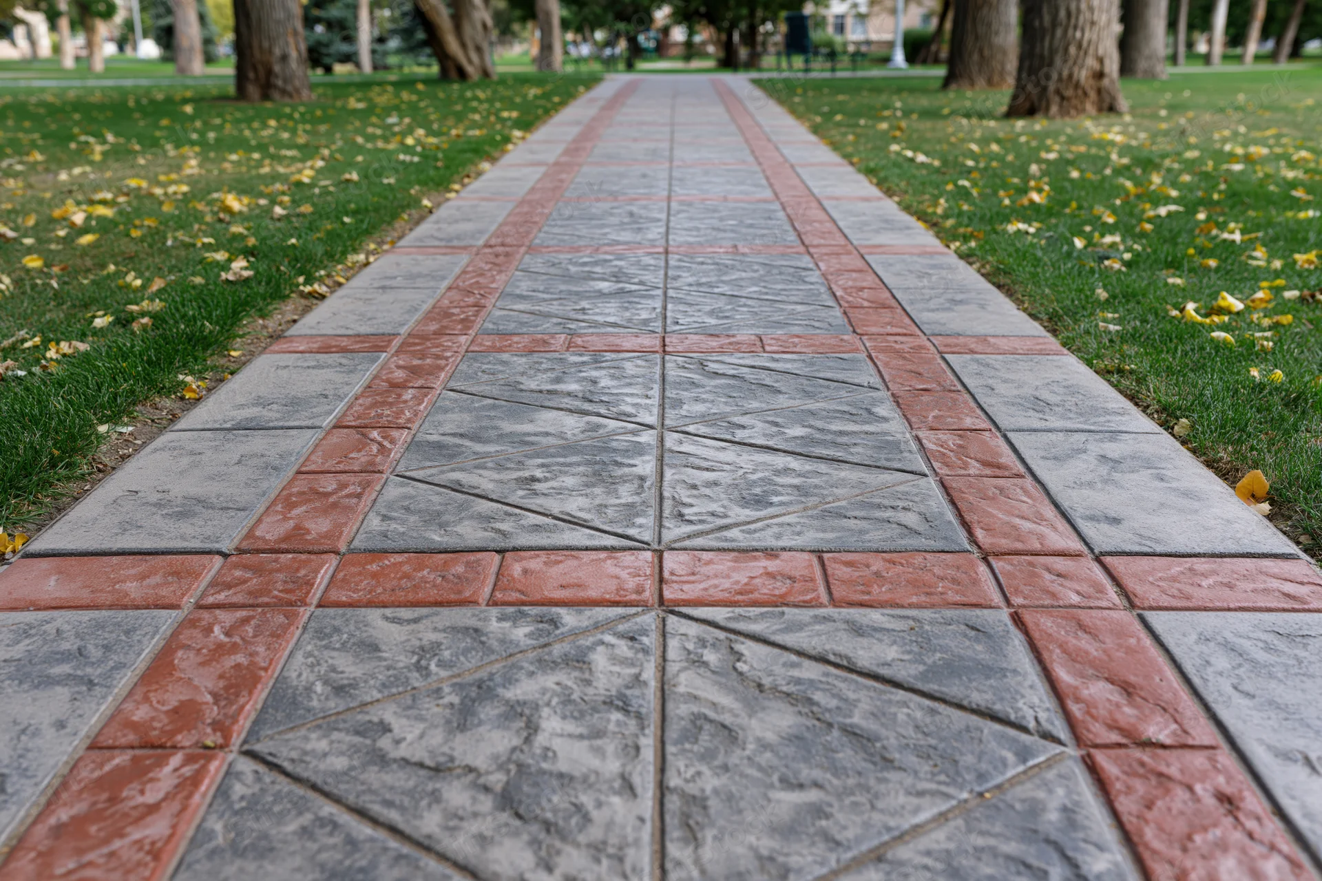 Stamped concrete pathway with red and gray geometric pattern in a public park surrounded by grass and trees with fallen autumn leaves