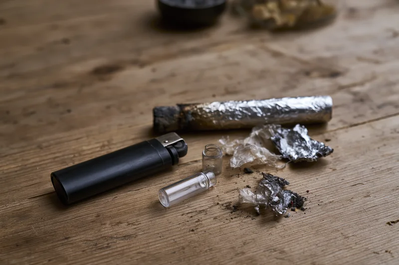 Drug paraphernalia including a burnt joint, foil, lighter and glass pipe arranged on a worn wooden surface
