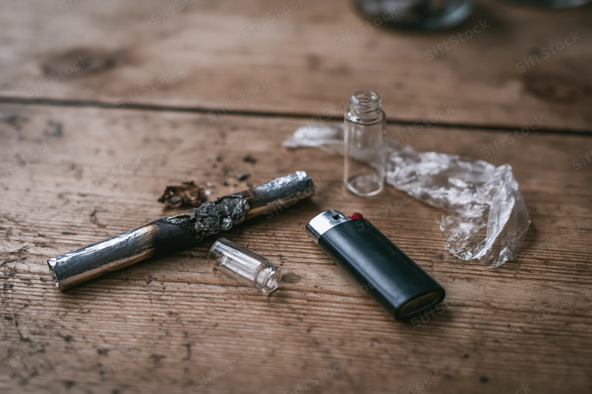 Burnt drug pipe and lighter with glass bottle and plastic wrap on worn wooden table