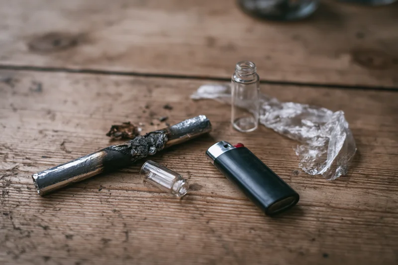 Burnt drug pipe and lighter with glass bottle and plastic wrap on worn wooden table