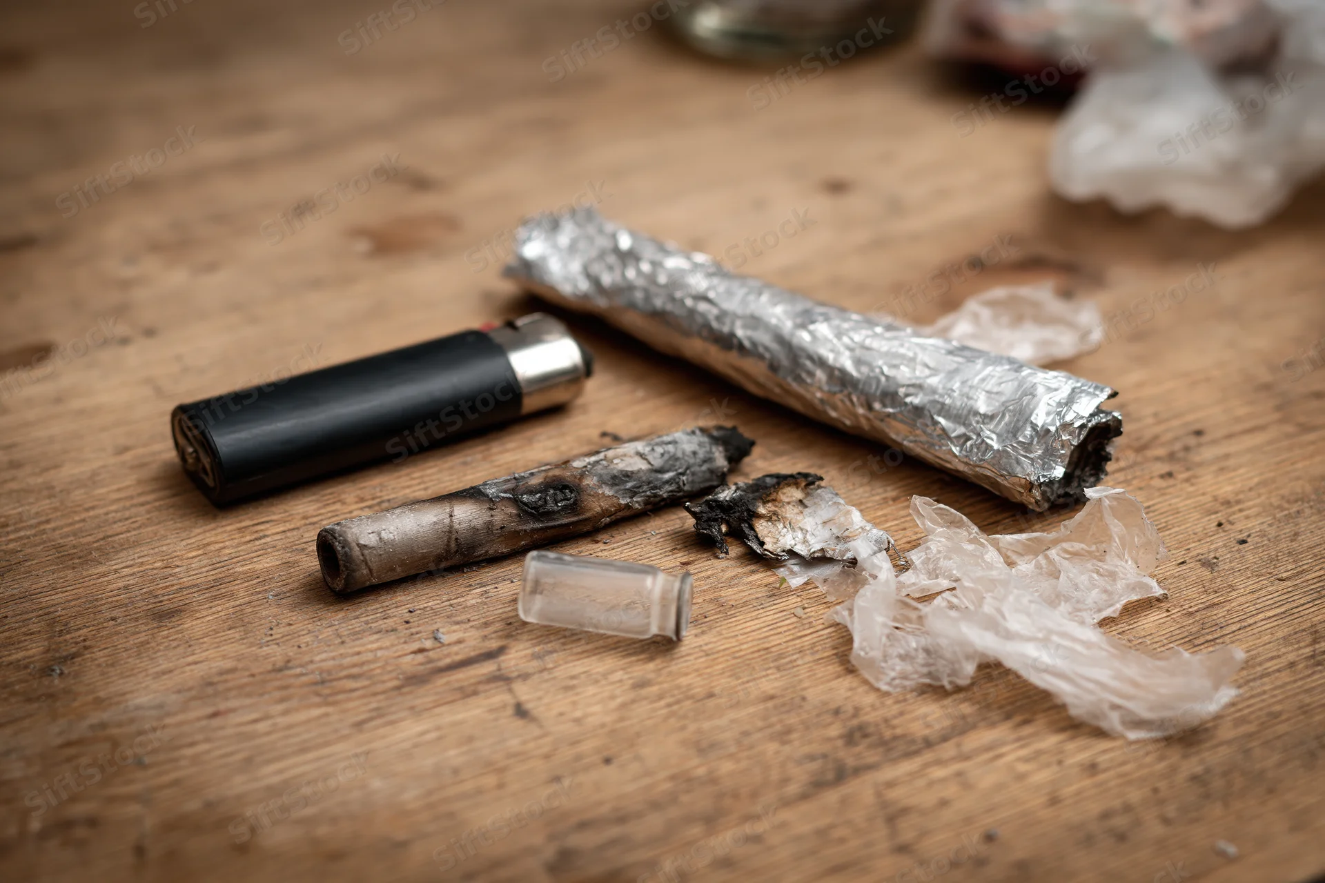 Drug paraphernalia including a burnt joint, foil-wrapped blunt, lighter, and ash scattered on a worn wooden surface