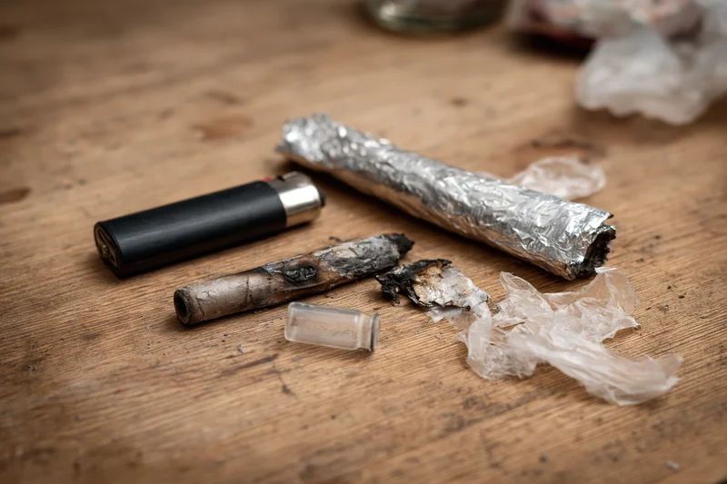 Drug paraphernalia including a burnt joint, foil-wrapped blunt, lighter, and ash scattered on a worn wooden surface