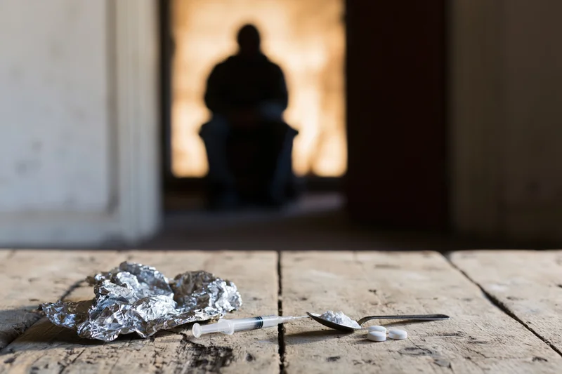 Drug paraphernalia on a worn wooden table with a silhouetted figure in the background