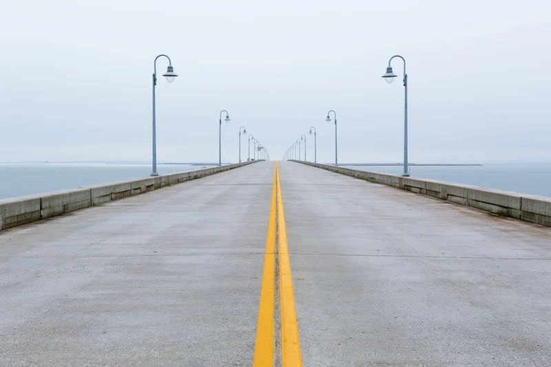 Empty concrete highway bridge stretching toward the horizon over water with yellow lane divider and lampposts on both sides