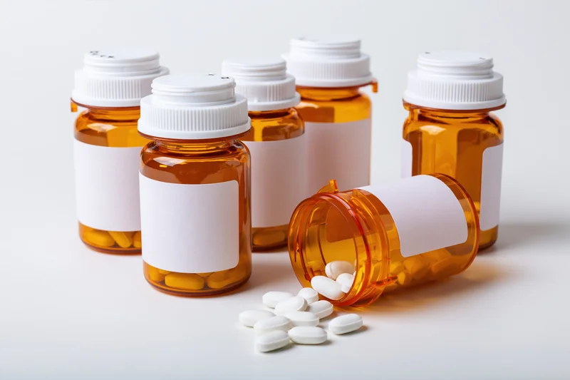 Several amber prescription bottles with white caps and blank labels, one tipped over with white pills spilled onto a white surface