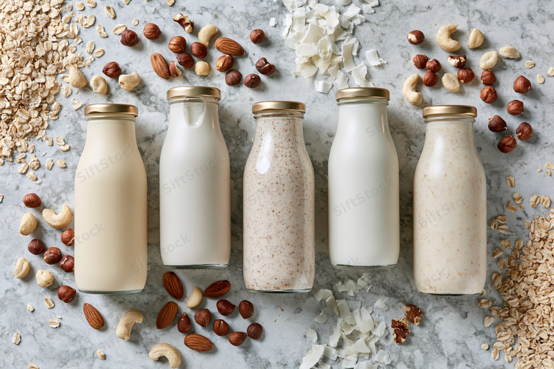 Five glass bottles of plant-based milk arranged in a row on a marble surface surrounded by scattered oats, nuts and coconut flakes