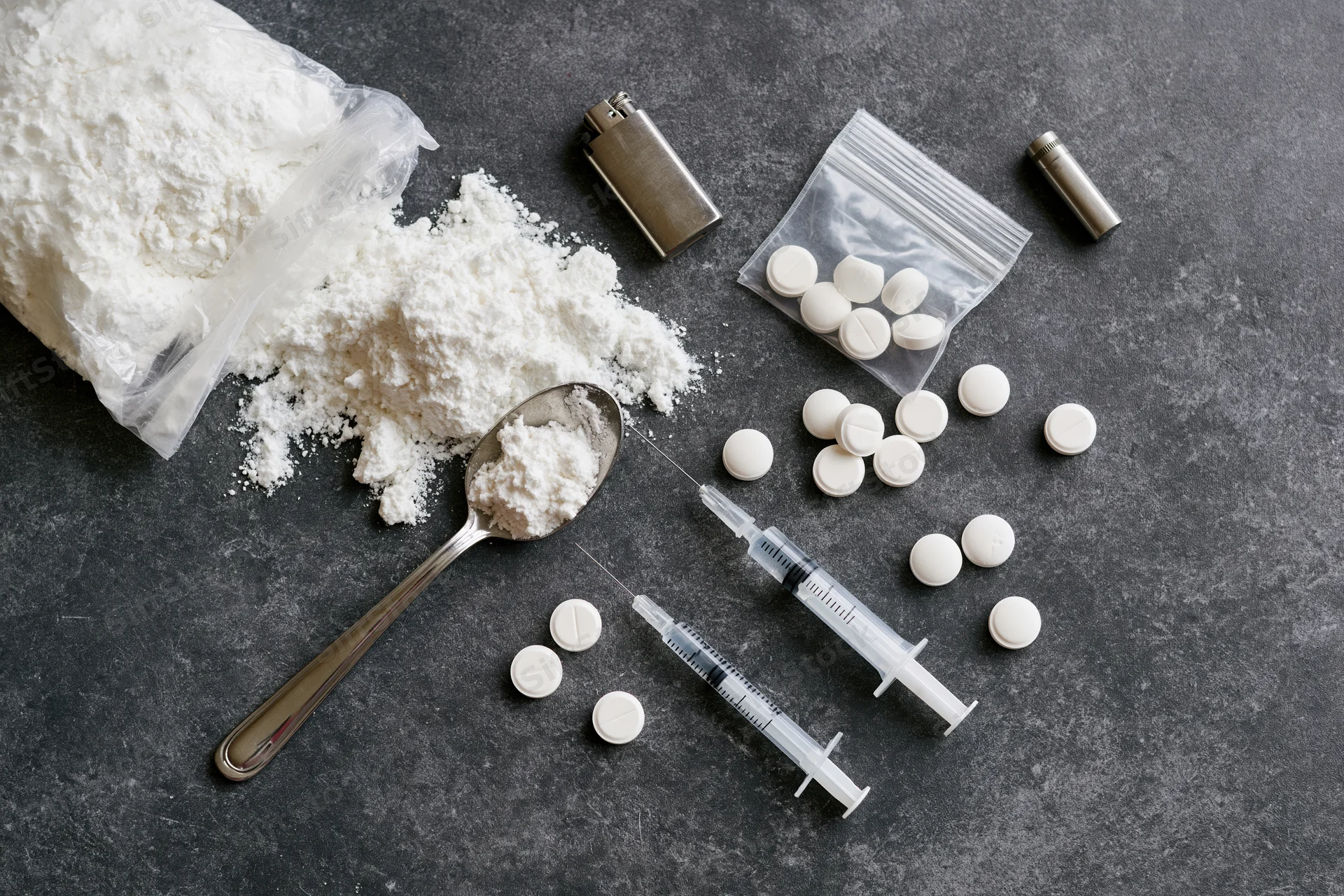 Drug paraphernalia arranged on dark textured surface including powder, syringes, pills, spoon, bag, lighter, and vial
