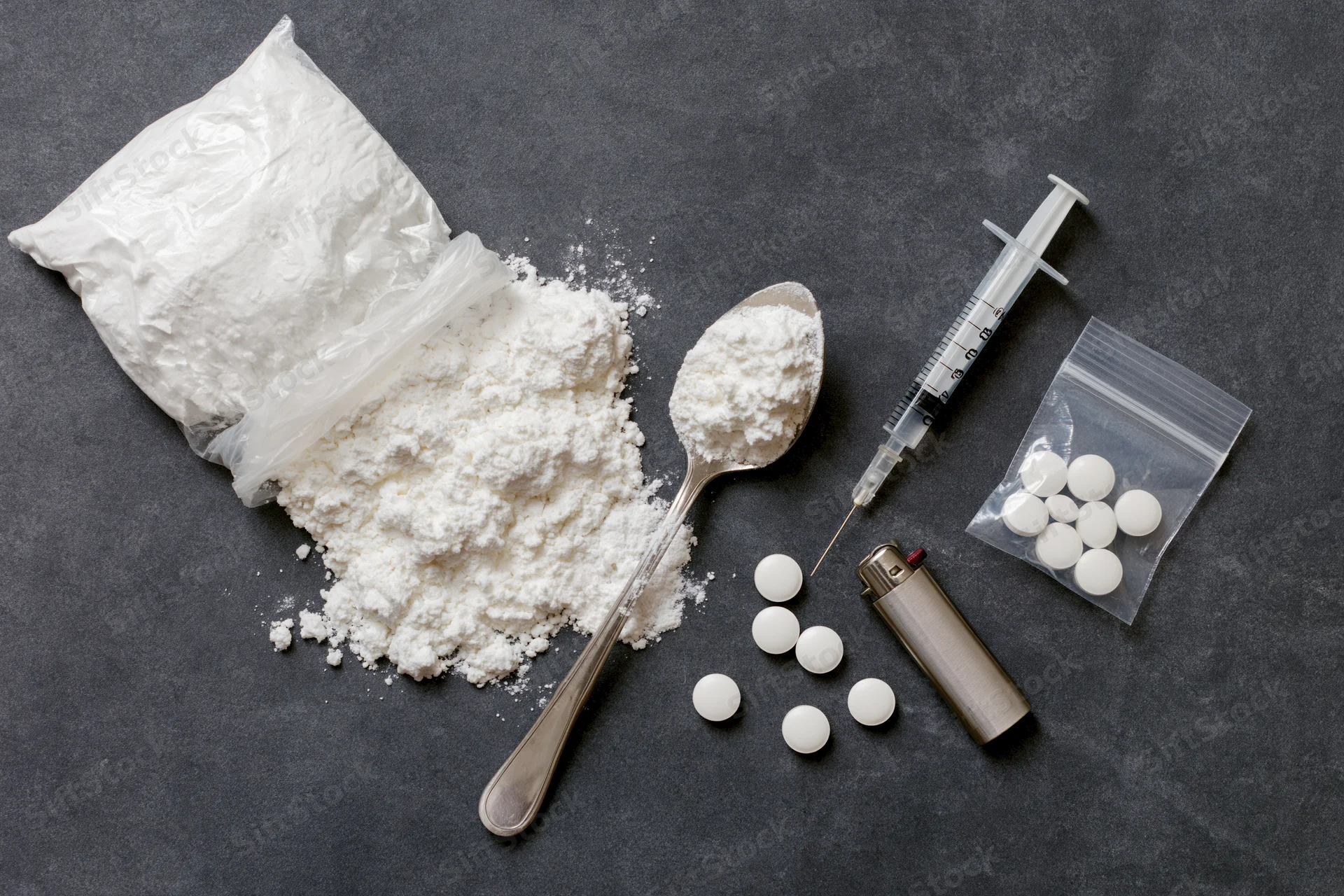 Drug paraphernalia including powder, syringe, spoon, pills, bag and lighter arranged on dark textured surface in flat lay overhead view