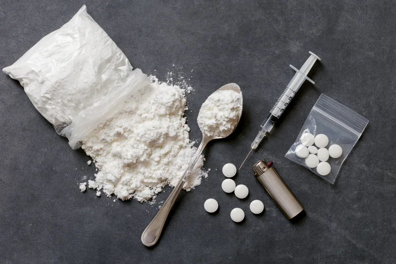 Drug paraphernalia including powder, syringe, spoon, pills, bag and lighter arranged on dark textured surface in flat lay overhead view