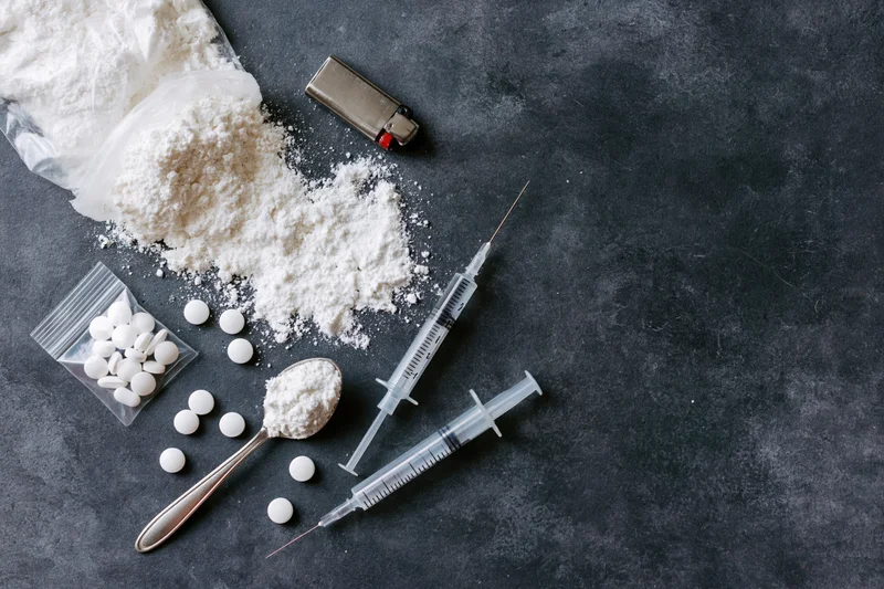 Drug paraphernalia including syringes, powder, pills, spoon and lighter arranged on dark textured surface
