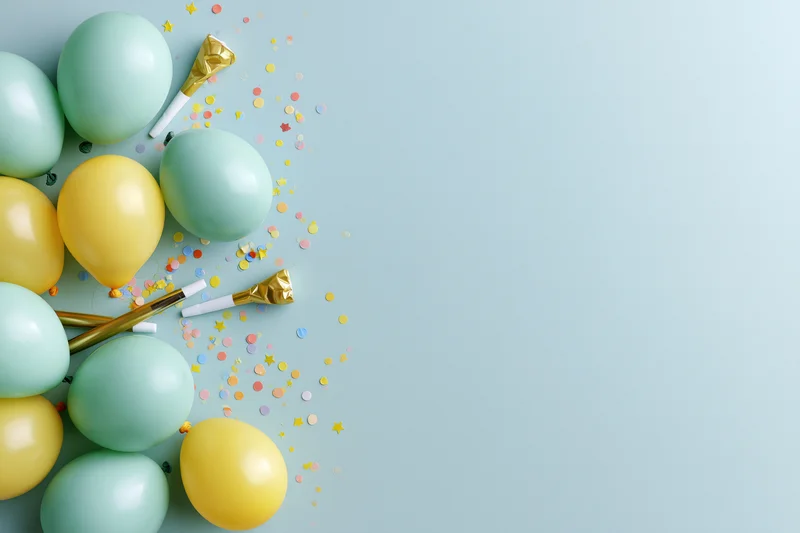 Mint green and yellow balloons with confetti and party blowers arranged in a flat lay on a light blue surface