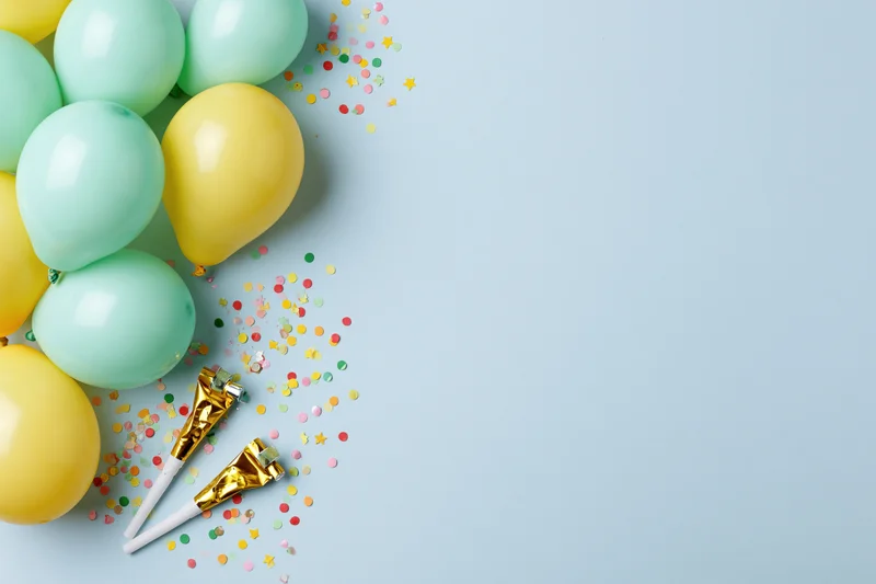 Mint green and yellow balloons with confetti and party horns arranged in a flat lay on a light blue surface