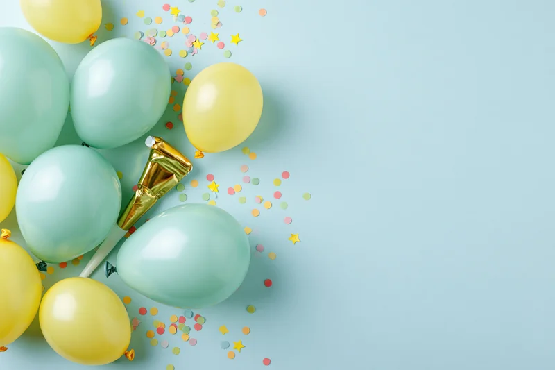 Mint green and yellow balloons with confetti and a gold party blower arranged in a flat lay on a light blue surface