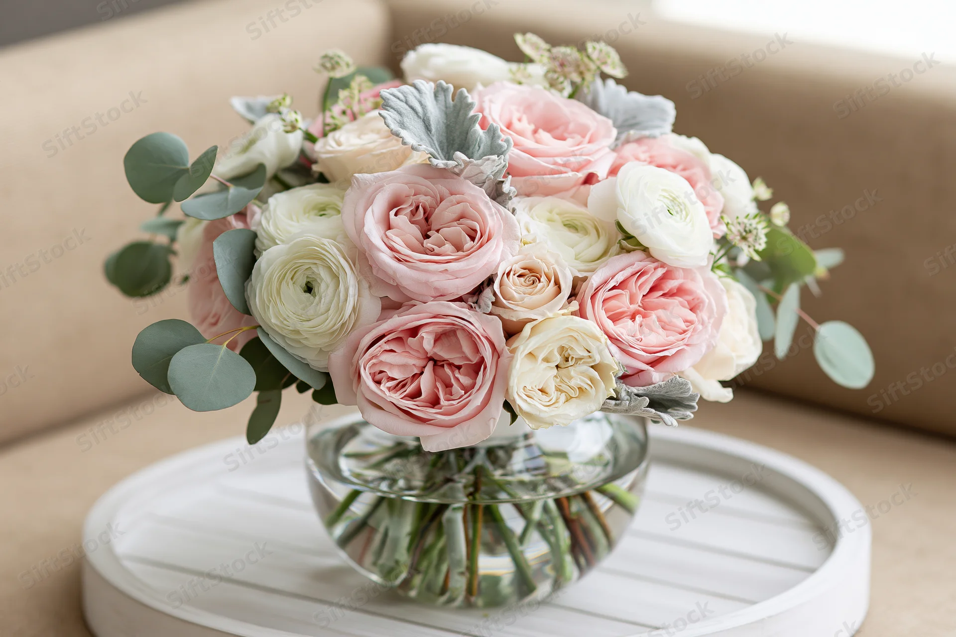Floral bouquet of pink roses and white ranunculus with eucalyptus and dusty miller in a glass vase on a white tray in a living room