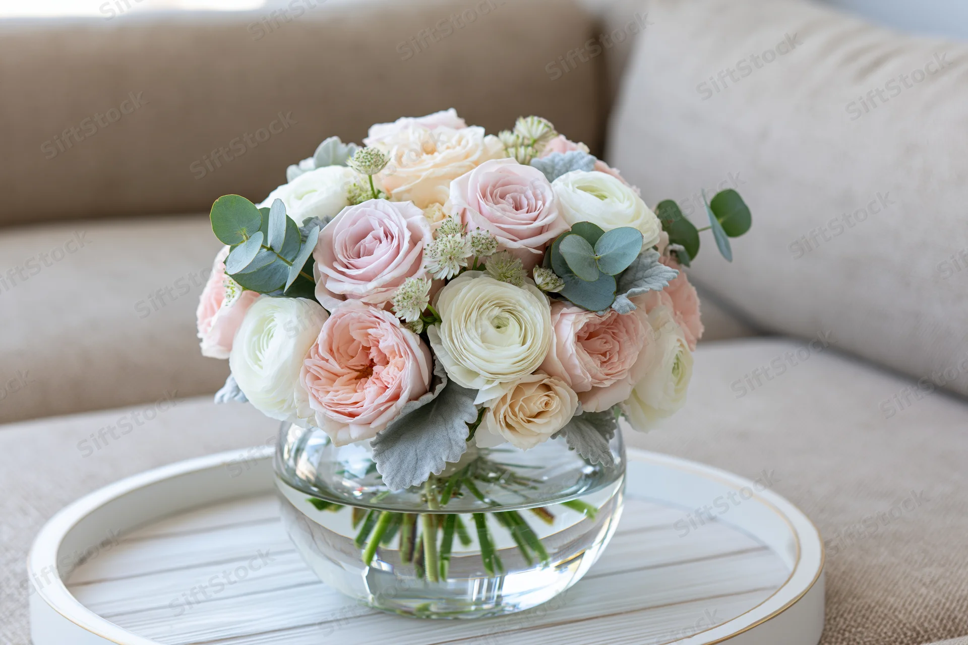 Floral bouquet of blush pink roses and white ranunculus in a glass vase on a white tray beside a beige sofa