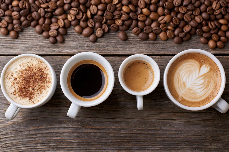 Four white ceramic coffee cups in a row on a dark wooden table with coffee beans above