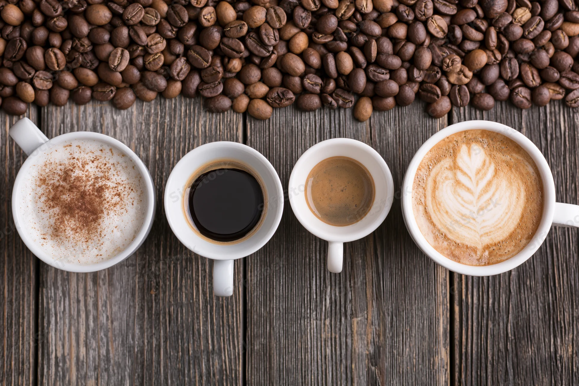 Four white ceramic coffee cups in a row with different coffee types on a dark wooden table, surrounded by roasted coffee beans
