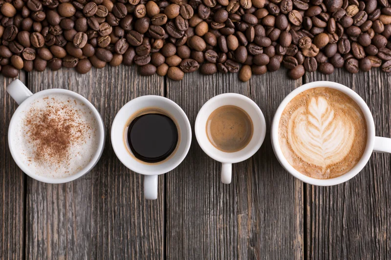 Four white ceramic coffee cups in a row with different coffee types on a dark wooden table, surrounded by roasted coffee beans