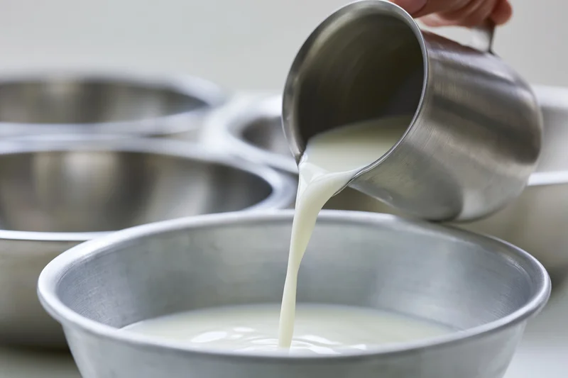 Milk being poured from a stainless steel pitcher into a metal bowl in a kitchen setting