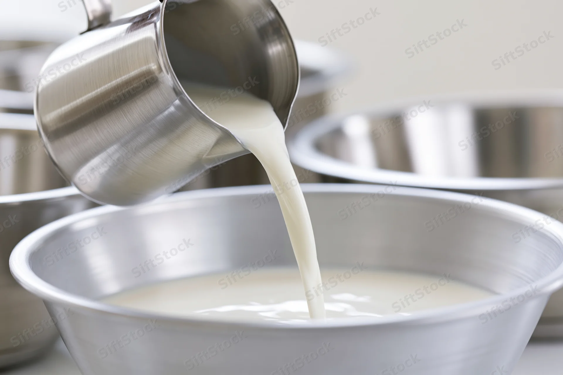 Milk being poured from a stainless steel pitcher into a metal bowl in a kitchen setting