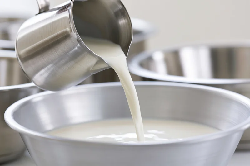 Milk being poured from a stainless steel pitcher into a metal bowl in a kitchen setting