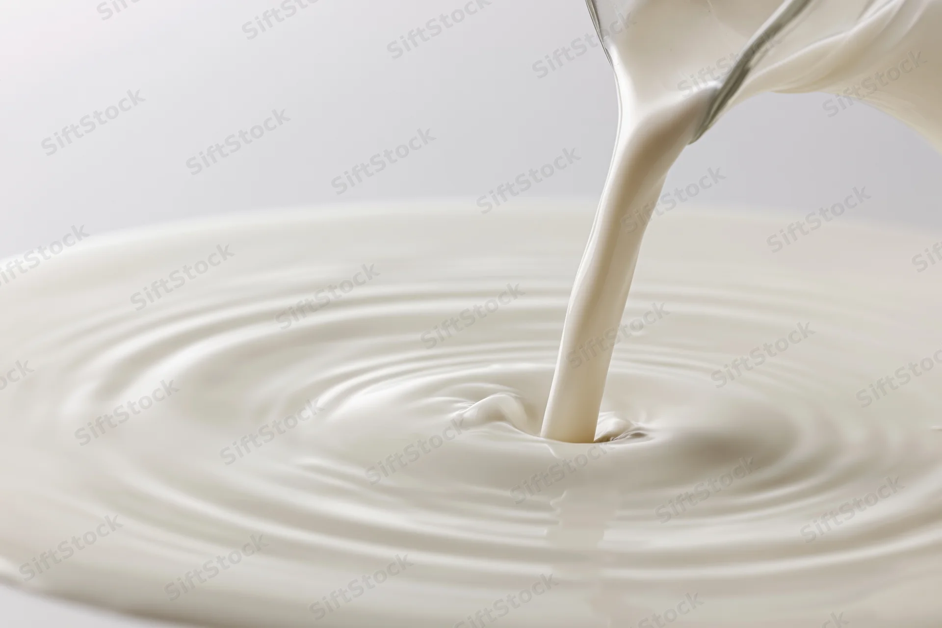 Milk pouring from a glass pitcher into a pool creating ripples on a white surface