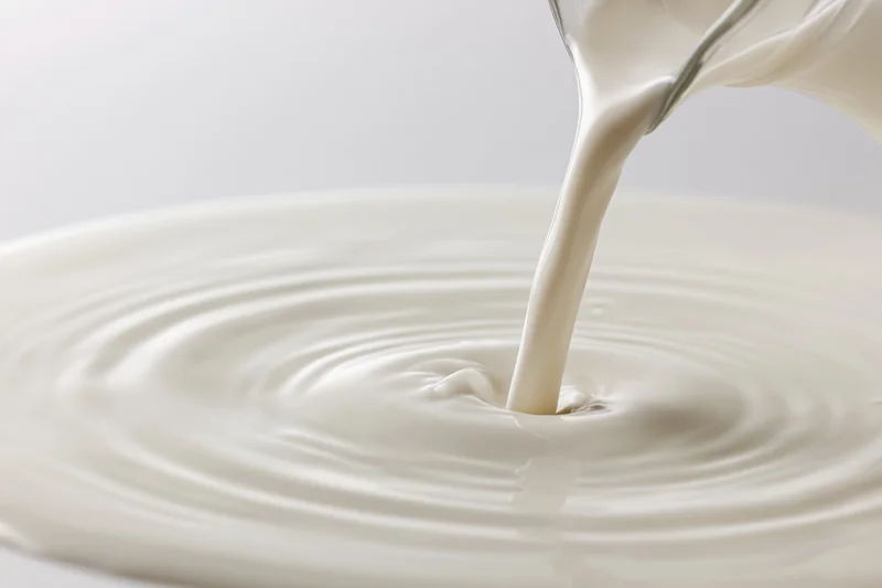 Milk pouring from a glass pitcher into a pool creating ripples on a white surface