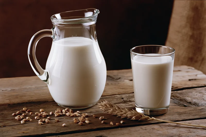 Glass pitcher and drinking glass filled with milk on a wooden table with oats and wheat stalks