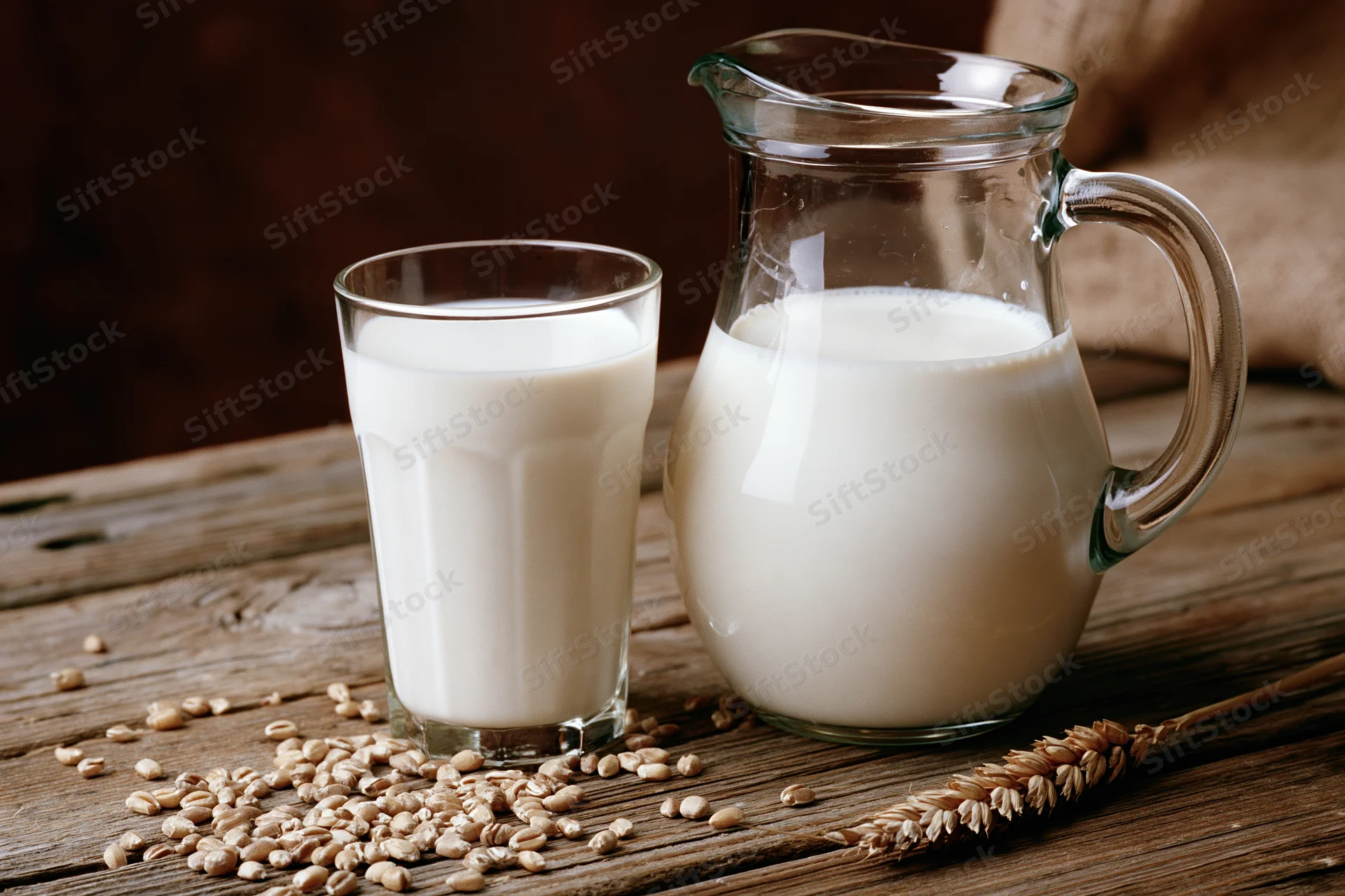 Glass of milk and pitcher on wooden table with scattered oats and wheat stalk