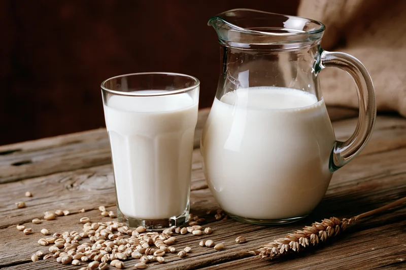 Glass of milk and pitcher on wooden table with scattered oats and wheat stalk