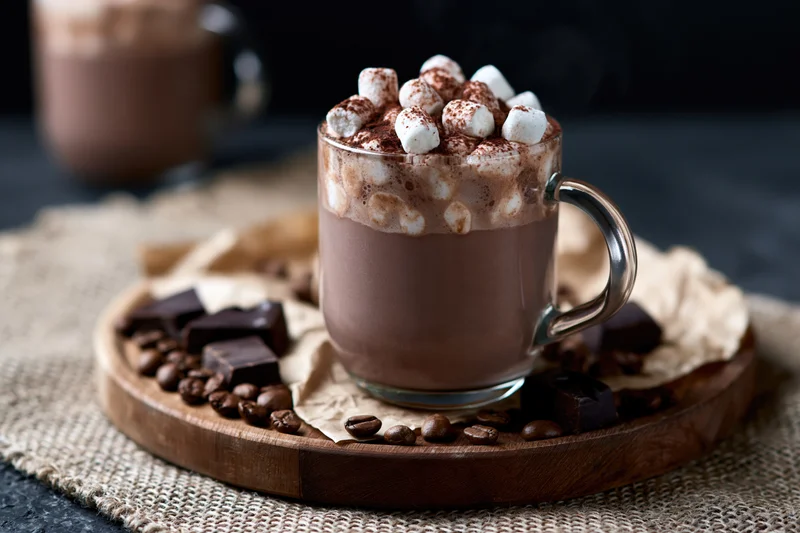 Glass mug of hot chocolate topped with mini marshmallows and cocoa powder on wooden tray with chocolate pieces and coffee beans
