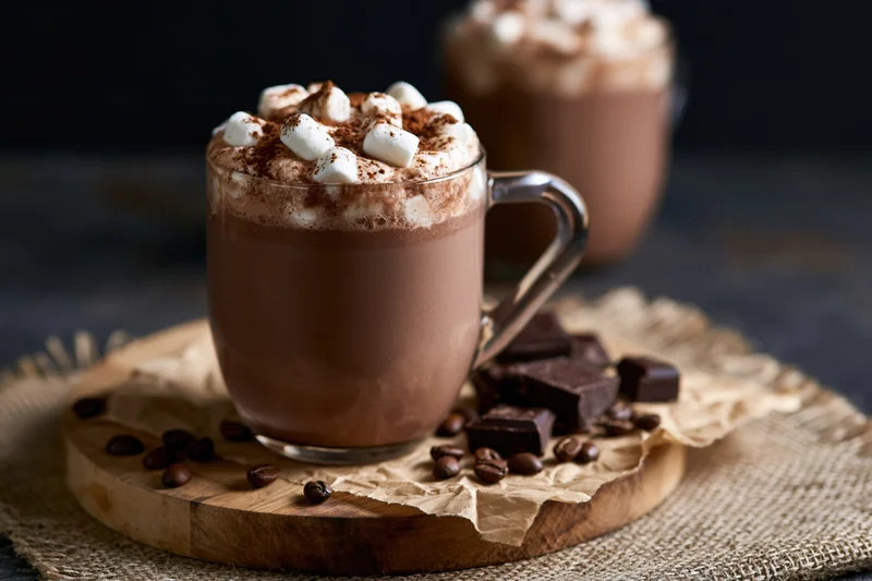 Glass mug of hot chocolate topped with mini marshmallows and cocoa powder on a wooden board with chocolate pieces and coffee beans