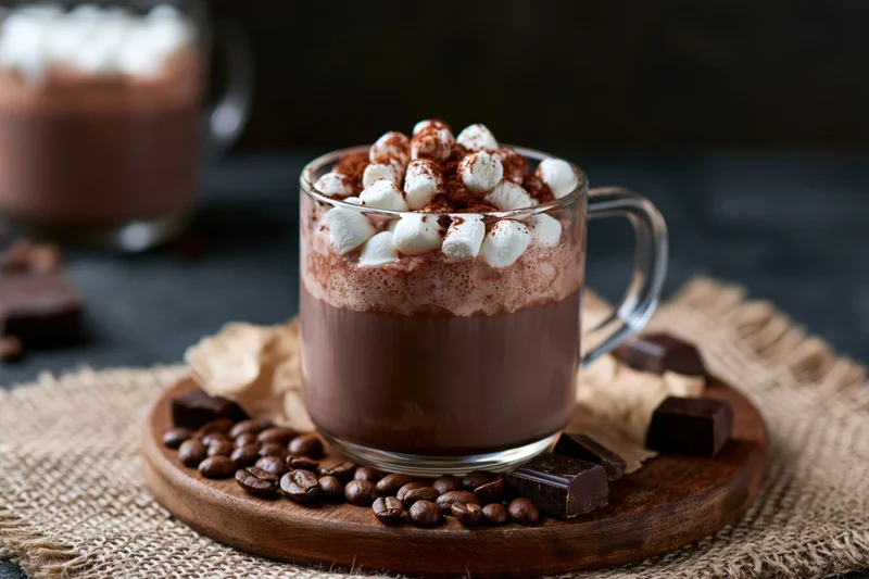 Glass mug of hot chocolate topped with mini marshmallows and cocoa powder on a wooden tray with chocolate pieces and coffee beans