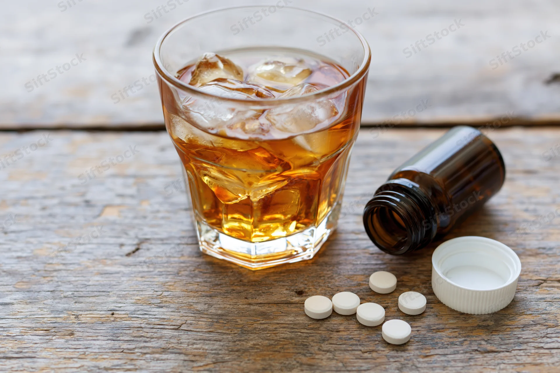 Glass of amber whiskey with ice and scattered pills on a worn wooden table