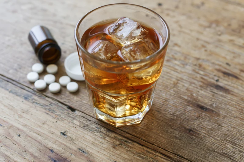 Glass of amber whiskey with ice cubes on a worn wooden table beside white pills and a small bottle cap