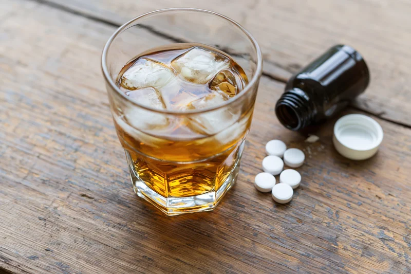 Glass of amber whiskey with ice and scattered pills beside a bottle on a worn wooden table