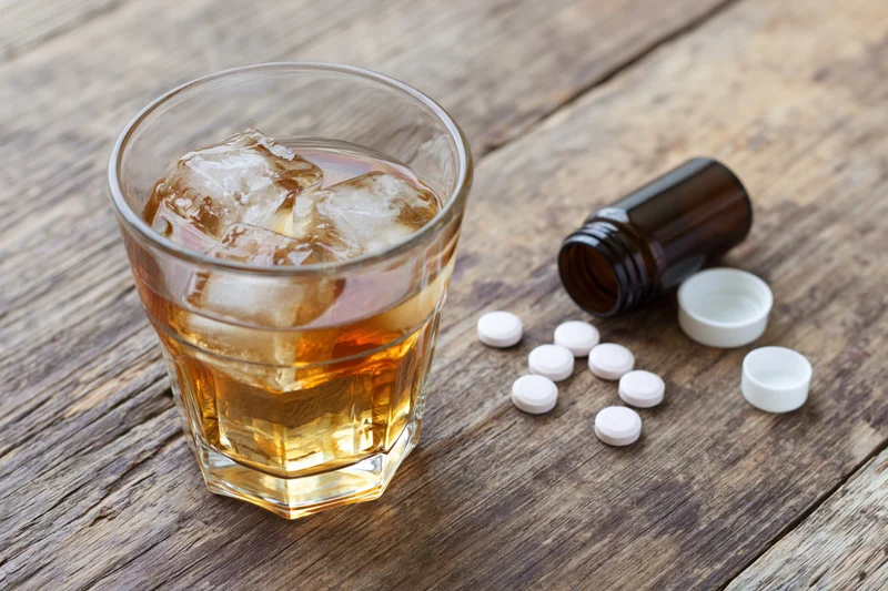 Glass of amber whiskey with ice and scattered white pills beside a bottle on a worn wooden table