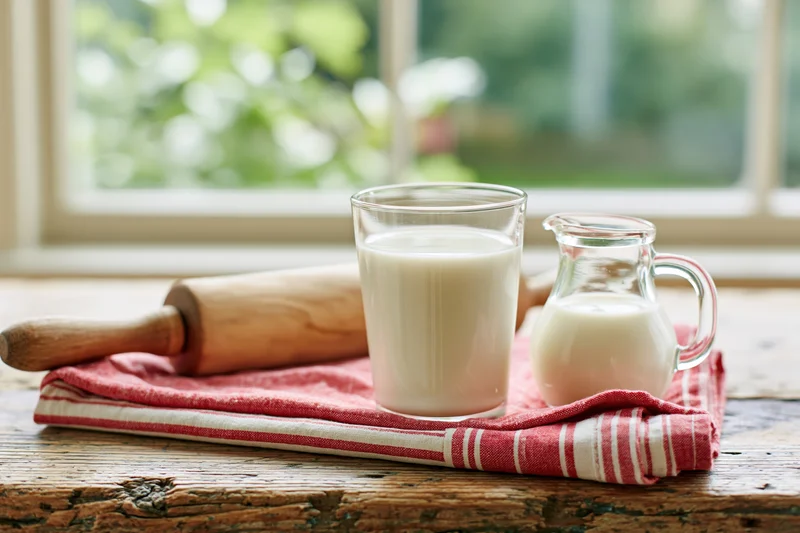 Glass of milk and small milk jug on a red striped towel with a wooden rolling pin on a rustic wooden table near a window