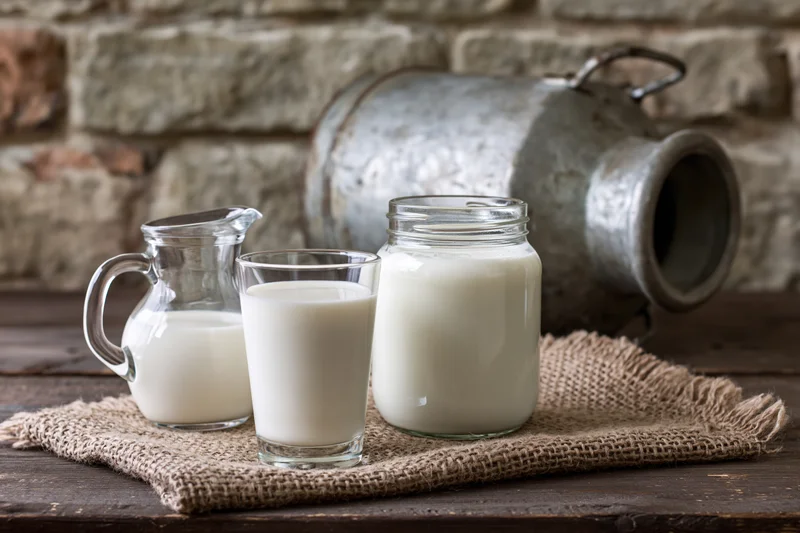 Glass of milk, small pitcher and jar filled with fresh dairy on wooden table with stone wall background