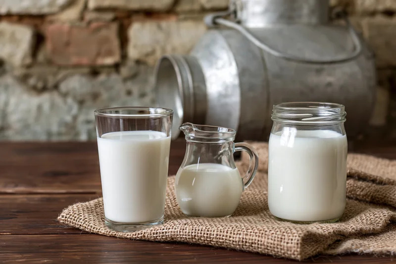 Glass of milk, small pitcher and jar filled with fresh milk on wooden table with burlap cloth and vintage milk can in background