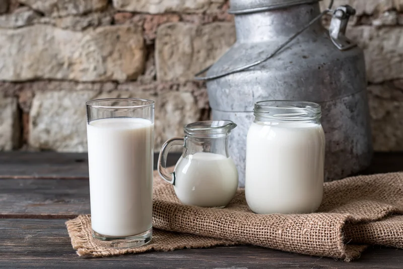 Glass of milk, small pitcher and jar filled with fresh milk on wooden table with stone wall background