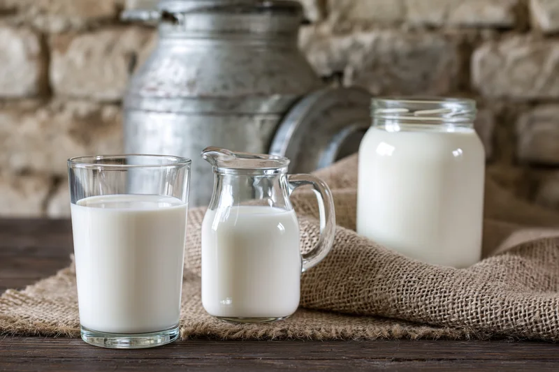 Glass of milk, small pitcher and jar filled with fresh milk on wooden table with burlap cloth and stone wall background