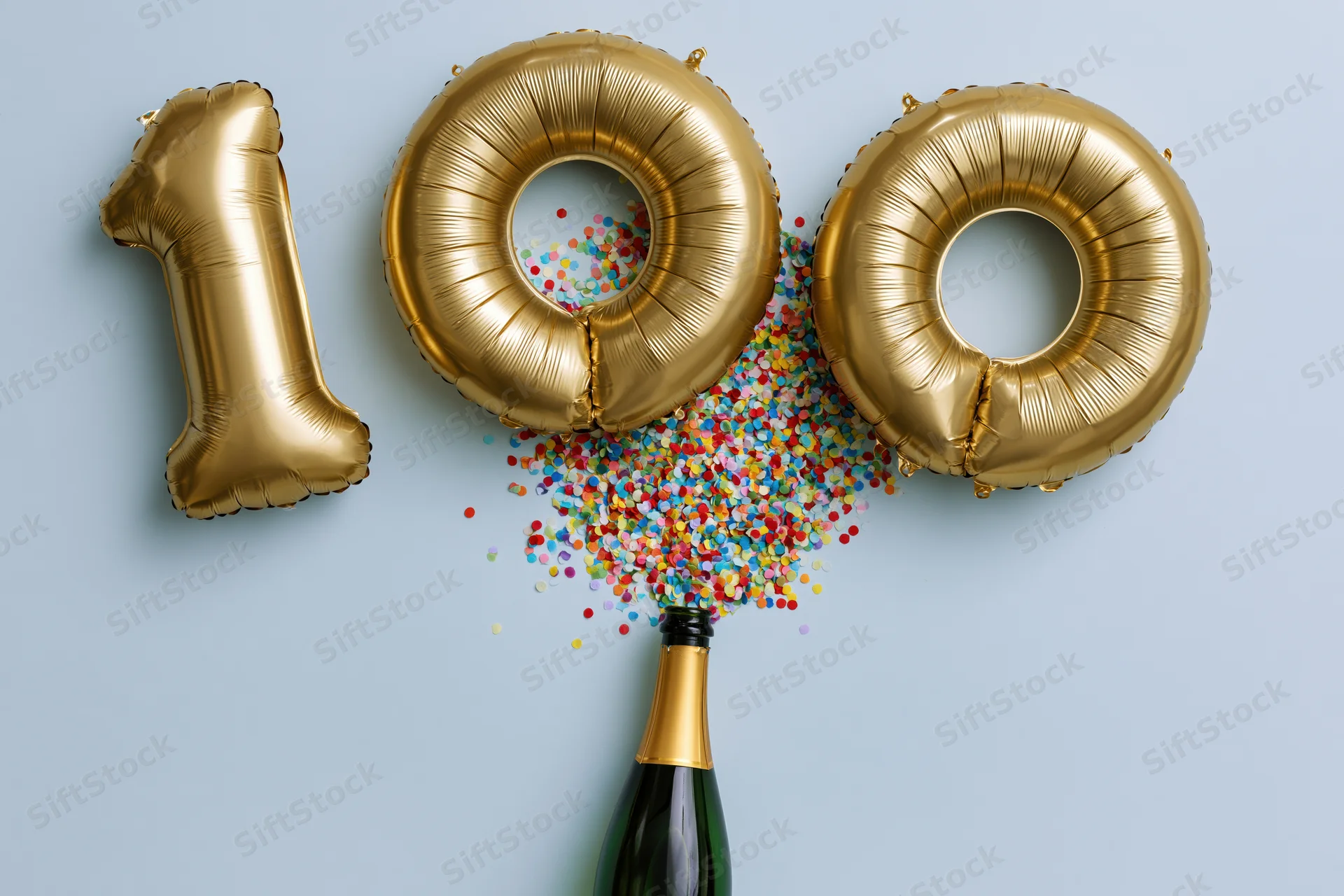 Gold foil number 100 balloons with confetti spilling from champagne bottle on light blue background