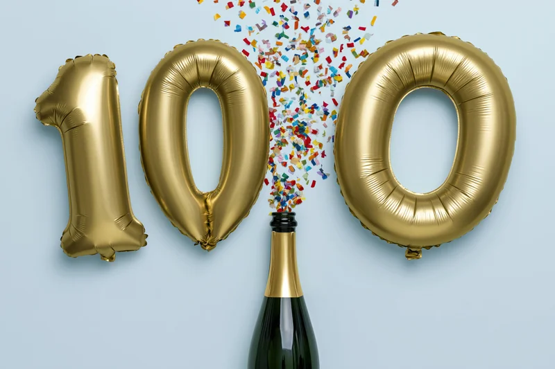 Gold foil number 100 balloons floating above a champagne bottle with confetti bursting from the top against a light blue background