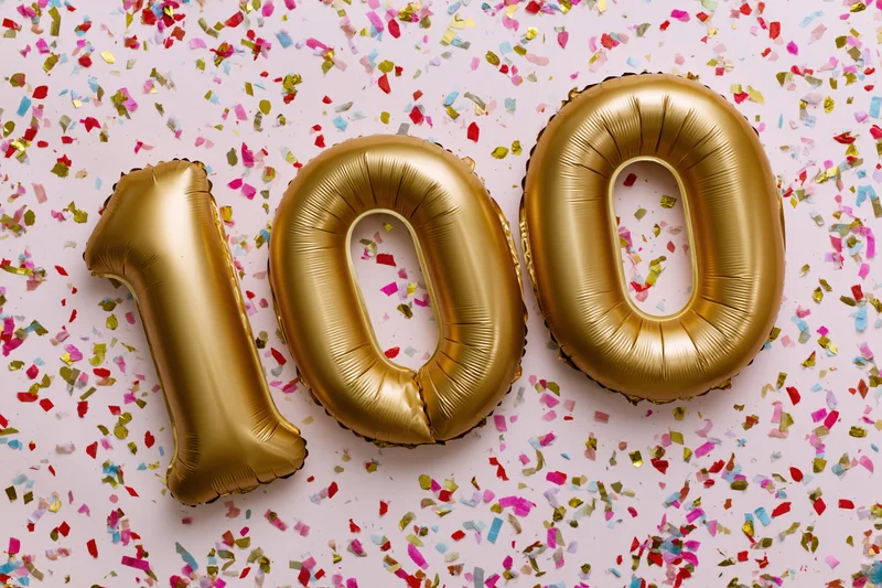 Gold foil number balloons spelling 100 on a pink surface scattered with colorful confetti
