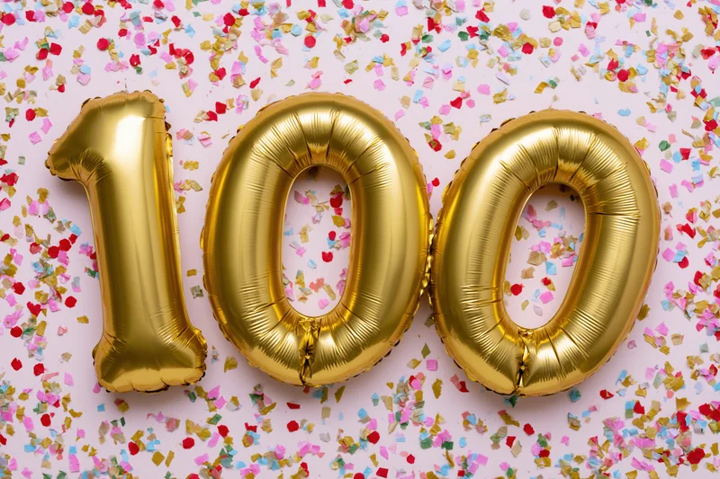Gold foil number balloons spelling 100 on a pale pink surface scattered with colorful confetti